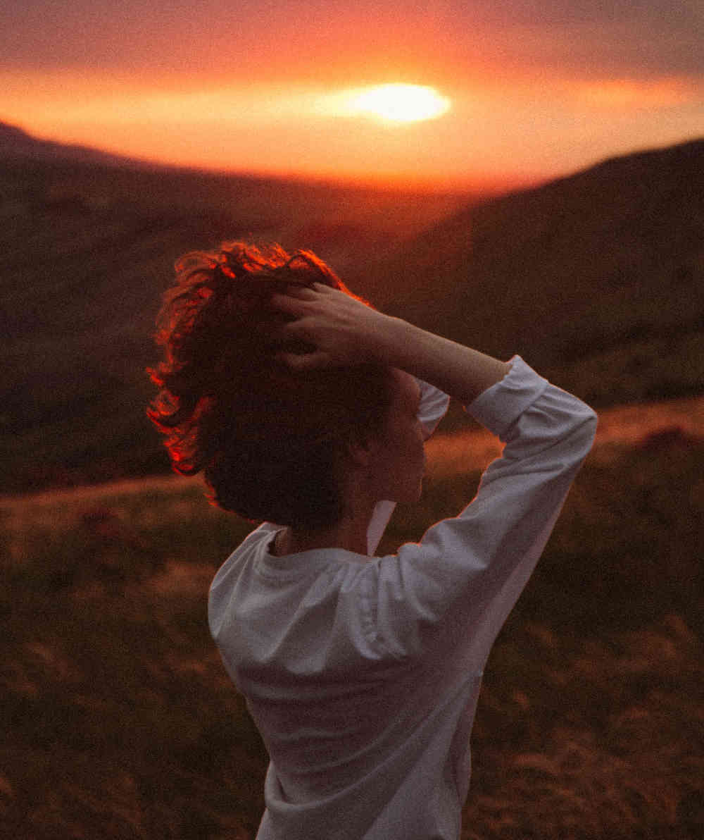 Woman in white shirt with hands to her head, standing in the foreground of hills, backlit by orange light of a setting sun.